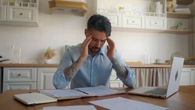 Overworked man feeling headache when working in home office in weekend, portrait. Exhausted freelancer sitting in home office in kitchen, rubbing temples after hard work overtime, man with laptop