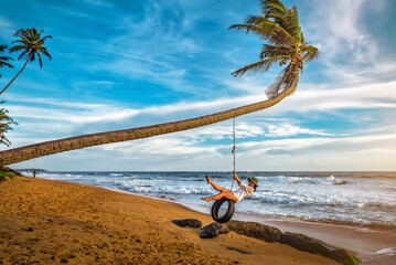 Tourist woman in hat having fun on tire swing on tropical beach - Powered by Adobe