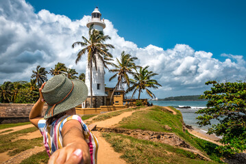Tourist woman in hat holds man hand an going to the lighthouse on tropical island. Fort Galle. Sri...