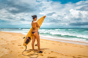 Young woman surfer holding her surfboard standing on the beach. Cloudy sky