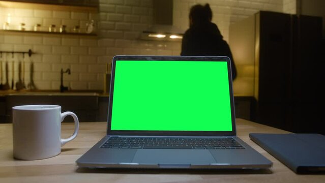 Laptop computer with mock up green screen chroma key display standing on the table. Man pressed to keyboard and dancing in the background in kitchen at night.