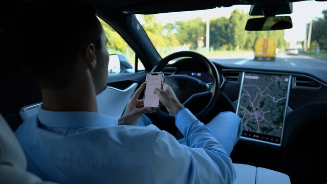 Successful businessman browsing modern smartphone while riding an autonomous self driving electric car. Male entrepreneur using phone during riding on electrical auto with autopilot at urban road