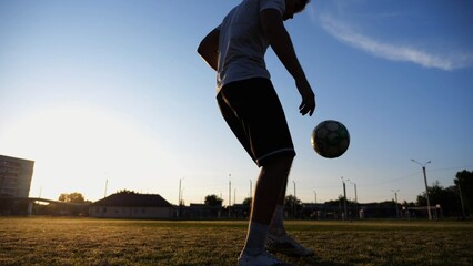 Professional footballer juggling soccer ball on stadium at sunset. Young man kicking ball at green field. Sportsman practicing tricks at meadow with sunlight at background. Freestyle football concept