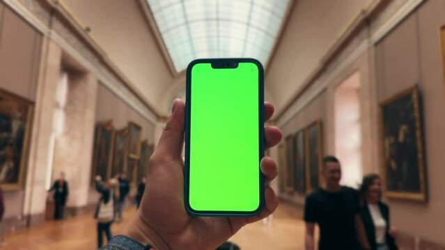Close up of woman's hand holds phone with green screen mock up. Blurred masterpiece art at background. Concept of digital app for museums and virtual excursion.