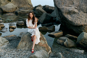 Woman in white dress sitting on ocean rock with red shoes, gazing at the horizon