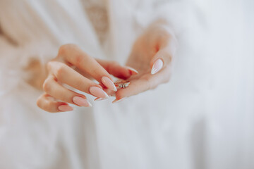 A woman is holding a ring in her hand. The ring is white and has a diamond on it. The woman is wearing a white robe and has long, painted nails. Concept of elegance and sophistication