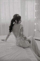 A woman in a wedding dress sits on a bed. She is wearing a tiara and has her hair in a ponytail