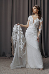 Fototapeta premium A woman in a white wedding dress is holding a white gown. The dress is long and has a lace design. The woman is wearing a tiara and has her hair in a bun. Concept of elegance and sophistication