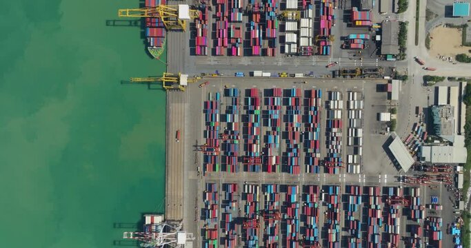 commercial port loading and unloading cargo from container ship import and export by crane for distributing goods by trailers transported to customers and dealers, aerial top down view 4k video