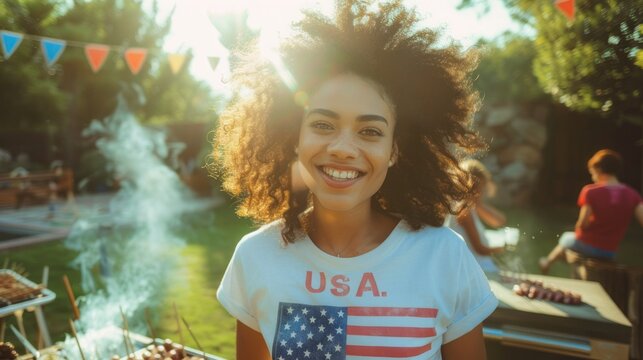 A young smiling American woman with an American flag t-shirt stands on the back door of house near a barbecue, the concept of celebrating independence day in America on the 4th of July with friends