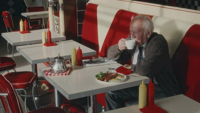 Side shot of good looking Caucasian man in advanced years sipping morning coffee while having breakfast in 1980s style diner