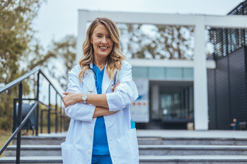 A smiling Caucasian woman in a blue medical scrub stands confidently outside a healthcare facility,...