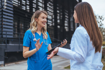 Two female healthcare workers engage in conversation outside a clinic, one in scrubs holding...