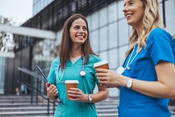 Two cheerful Caucasian female nurses enjoy a well-deserved coffee break outside a medical facility,...