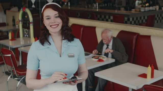 Medium portrait of pretty Caucasian waitress with red hair wearing light blue uniform and apron, writing down orders and looking at camera with smile in 1980s style American diner