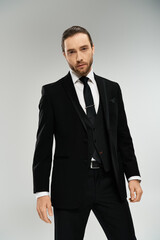 A bearded businessman exudes confidence and elegance as he poses in a suit and tie against a grey studio backdrop.