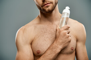 A handsome, shirtless man with a beard holds a bottle of facial cleanser