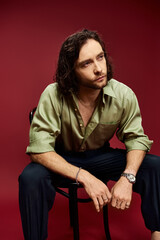 A handsome man in glasses and a green silk shirt sits serenely on a chair against a vibrant red background.