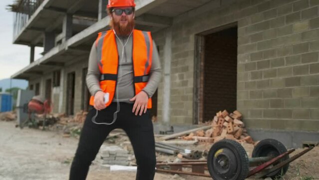 Incendiary dance of a charismatic builder with a red beard listening to music in phone headphones while dancing in an orange reflective vest and hard hat on construction site.