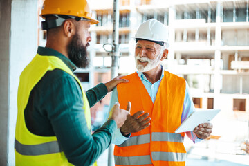 A male construction worker in a green jacket converses with a smiling male site manager in an...