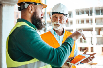 A younger worker in a green vest and hard hat shows a tablet to an older site manager in white,...