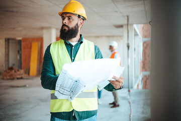 A bearded man wearing a yellow hard hat and reflective vest studies architectural plans at a...