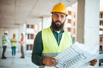 A focused male construction worker with a beard examines construction plans, wearing a yellow hard...