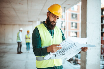 A bearded male construction worker in a yellow hard hat inspects architectural plans at a bustling...