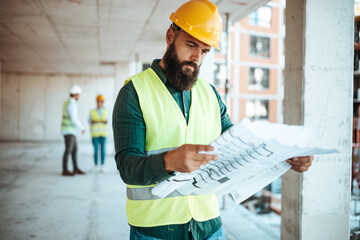 A bearded male construction worker in a yellow helmet and high-visibility vest examines blueprints...
