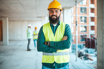 A male construction worker with a beard stands confidently at a construction site, arms crossed,...