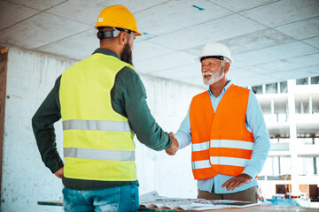 A bearded construction worker in a yellow hard hat greets a senior manager wearing a white helmet with a warm handshake, surrounded by blueprints and the structure of a building under construction.