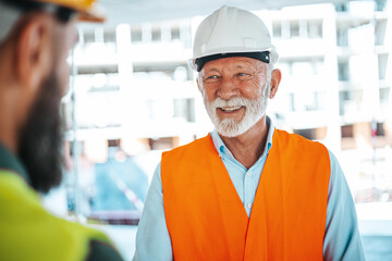 A smiling senior construction manager in a white helmet and orange vest shaking hands with a...
