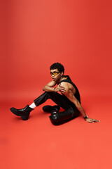 Handsome African American man in black leather pants sitting on a red background.