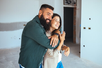 A joyful man and woman share a hug, surrounded by the bare walls of their newly acquired apartment,...