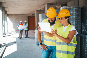 A male and female engineer in safety gear engage with digital blueprints on a tablet, while...
