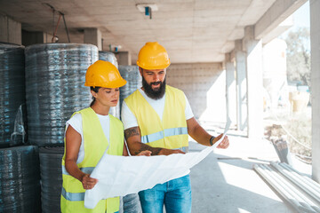A man and woman in hard hats and reflective vests discuss plans at a building site, exemplifying teamwork in the construction industry.