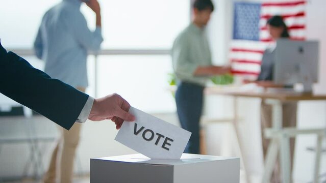 Close up of hand putting slip of paper with "Vote" written on it into ballot box at polling station for American election with voters - shot in slow motion