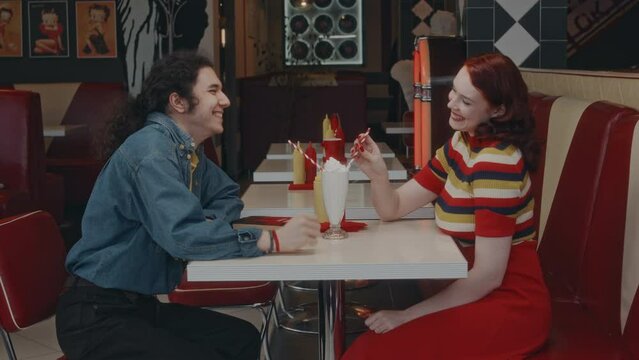 Side medium long shot of smiling Caucasian boyfriend and girlfriend drinking same milkshake in 1980s style diner, holding hands and flirting while on date