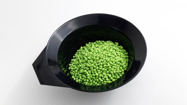 Light green masterbatch granules are poured from a scoop into a black measuring cup on a white background, this polymer is a colorant for products in the plastics industry.