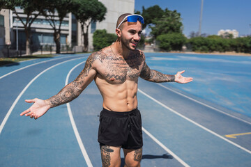 Chico joven musculoso y tatuado posando en pista de atletismo con ropa deportiva
