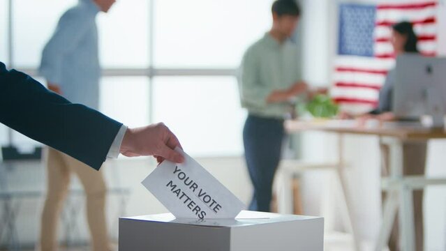 Close up of hand putting slip of paper with "Your Vote Counts" written on it into ballot box at polling station for American election with voters - shot in slow motion