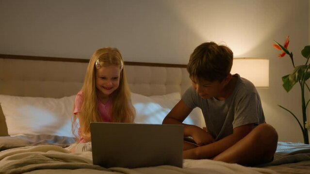 Happy family Caucasian children daughter son little kids watching movie video online Internet cartoon siblings boy girl using computer laptop bed night learning study distant education indoors bedroom
