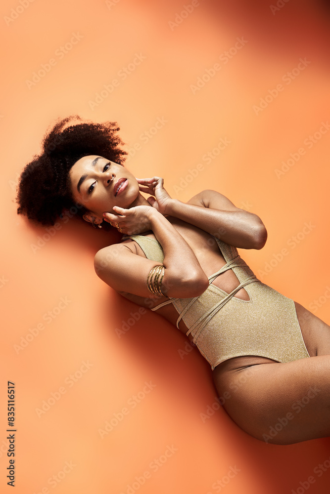 Wall mural stylish african american woman sunbathing in trendy swimsuit on bright orange background.