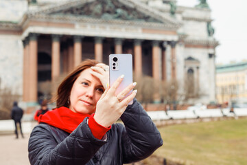 young woman looks at her smartphone as if in mirror and corrects her makeup on European city Saint...
