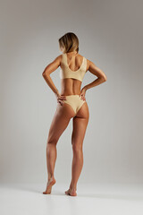 Rear view photo of young beautiful woman with slim toned body posing in beige lingerie against grey studio background. Concept of beauty and fashion, female health, anti-cellulite, dieting. Ad