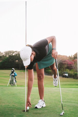 Obraz premium A young woman on a golf course at a country club bent down in front of the hole and reached out to reach the ball from below. Hitting the target and moving to another field. Professional golfer.
