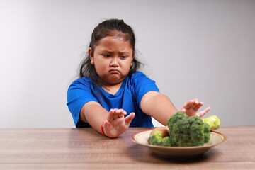 Unhappy asian little girl refusing to eat vegetables at table in room