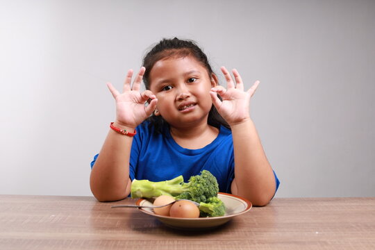 Happy cute asian little girl showing okay hand gesture with vegetables on the table. Healthy food concept