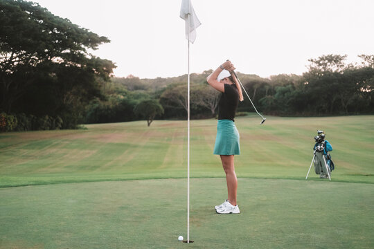 A young Caucasian woman in a sports skirt is a beginner golfer, she is practicing on the green lawn of a country club. The woman misses the hole and is upset, holding her head with a club in her hands