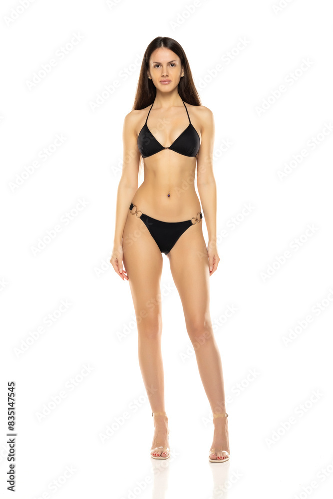 Wall mural young brunette woman in bikini swimsuit posing on a white studio background. front view